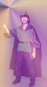 A slender, bearded young man dressed in green and violet tunic, cape and cap holds aloft a sword from which a soft, multicolored light eminates. [Thom about to head off to the Renaissance Faire]